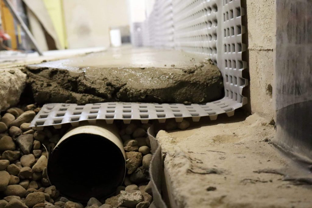 Close-up of interior French drain floor edging and drain tile installed beside a basement foundation wall.